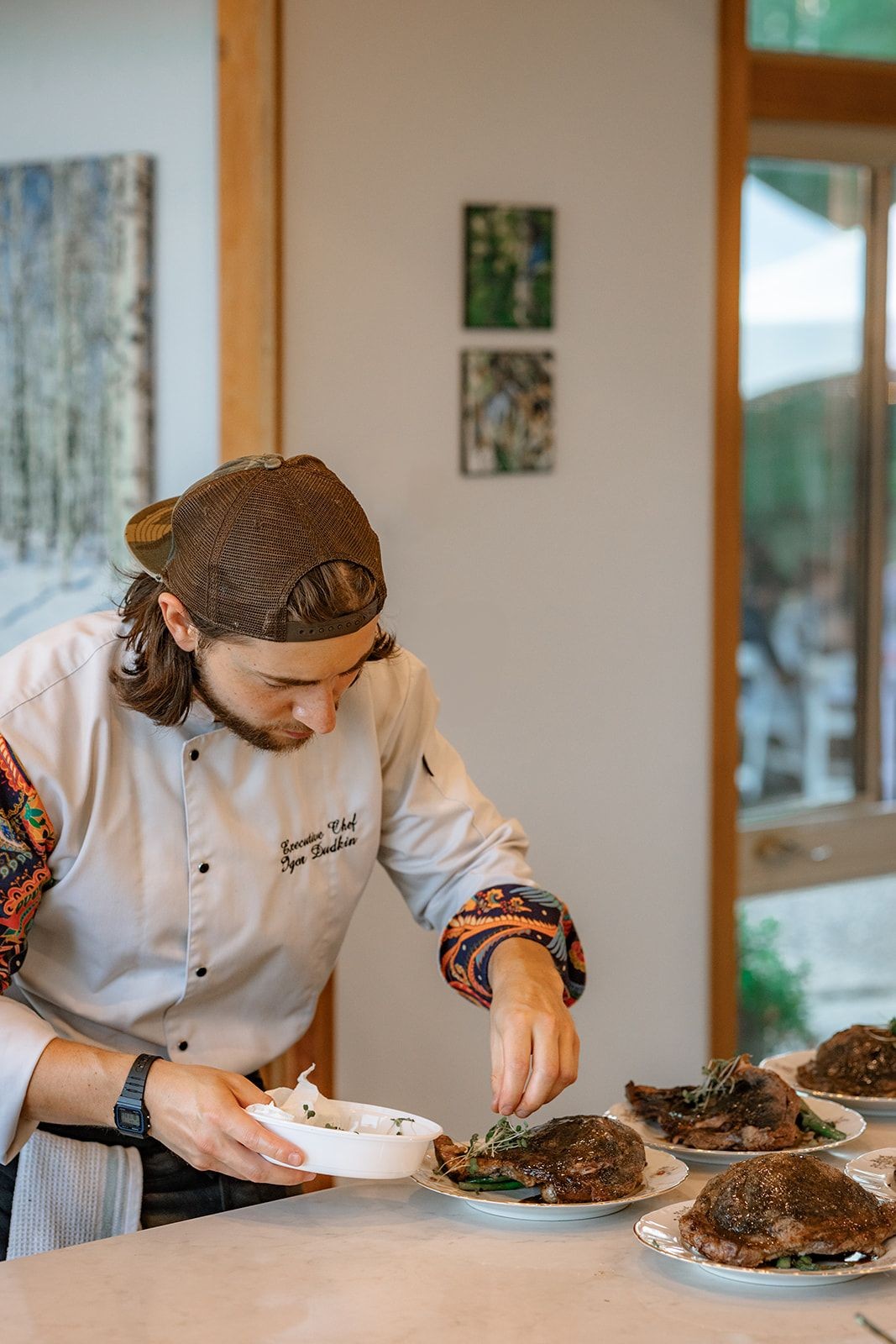 Chef in patterned uniform garnishes plates with microgreens in a kitchen setting.